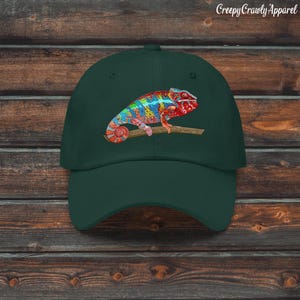 Könnte beinhalten: Dunkelgrüne Baseballkappe mit einem farbenfrohen Chamäleon-Design. Das Chamäleon sitzt auf einem Ast und hat ein lebendiges Muster aus Rot, Blau, Gelb und Grün. Der Text "Creepy Crawly Apparel" ist oben rechts aufgedruckt.