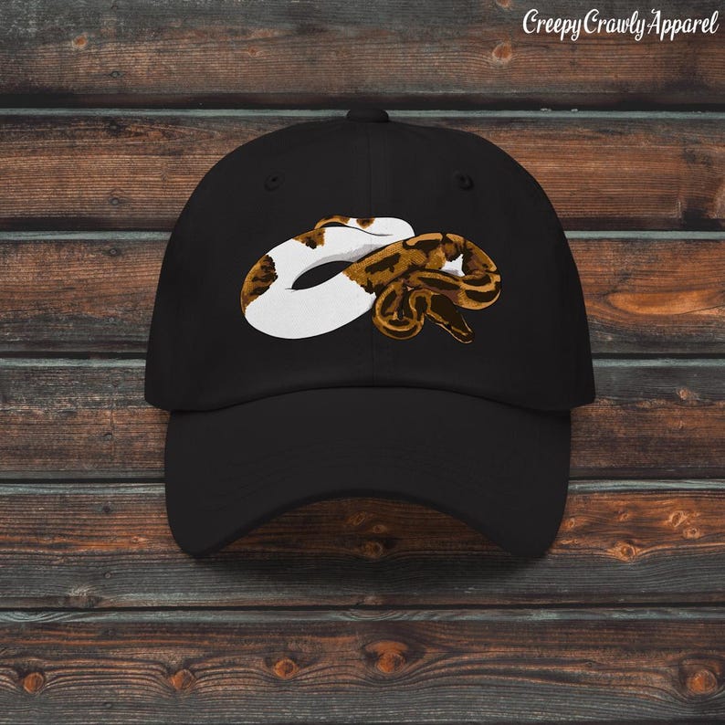 May include: Black baseball cap featuring a graphic of a snake in white, brown, and tan. The text "Creepy Crawly Apparel" is printed in white on the top right of the cap. The cap is set against a wooden background.