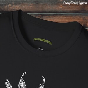Op de afbeelding: Zwart T-shirt met een witte afbeelding van een griezelig kruipend insect. Het shirt heeft een label met de tekst "CreepyCrawlyApparel" en "100% katoen".