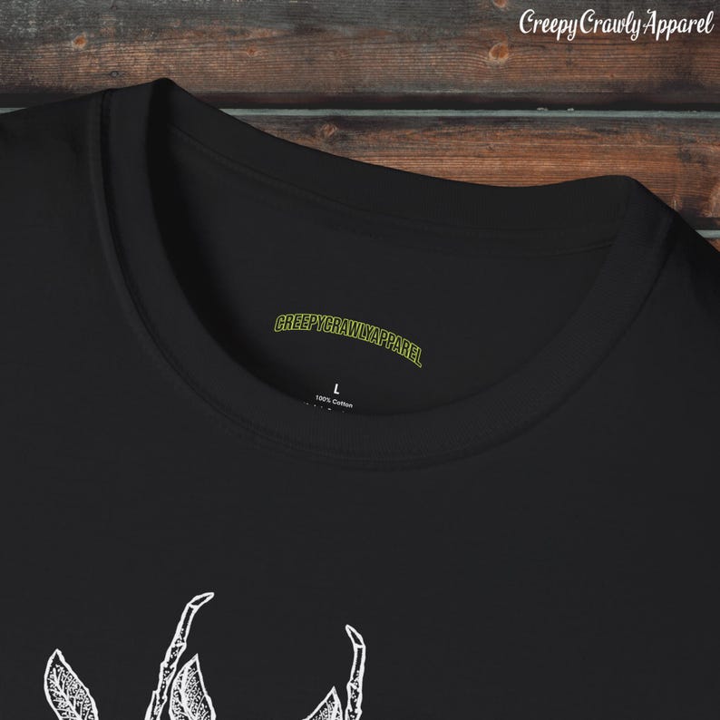 Op de afbeelding: Zwart T-shirt met een witte afbeelding van een griezelig kruipend insect. Het shirt heeft een label met de tekst "CreepyCrawlyApparel" en "100% katoen".