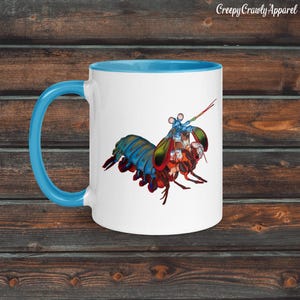 May include: White ceramic mug with a blue handle and interior. The mug features a colorful illustration of a mantis shrimp with a small figure riding on its back. The text "Creepy Crawly Apparel" is printed on the top right.