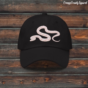 Könnte beinhalten: Eine schwarze Baseballkappe mit einer hellrosa Grafik einer zusammengerollten Schlange in der Mitte der Vorderseite. Das Design zeigt die glatte, gewundene Silhouette einer Schlange in einem sanften Pastellton auf dem dunklen Stoff.