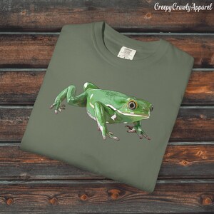 May include: An olive green t-shirt with a detailed illustration of a bright green frog with white markings. The frog is the main subject, set against the plain shirt. The text "Creepy Crawly Apparel" is in the top right corner.