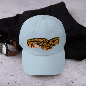 May include: A light blue baseball cap featuring a detailed illustration of a ball python. The snake is depicted in shades of brown, black, and yellow. The cap is a classic six-panel design with a curved brim.