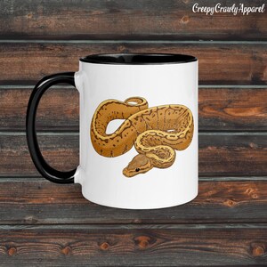 Può includere: Tazza in ceramica bianca con manico e bordo neri, con un'illustrazione dettagliata di un serpente marrone e beige. La tazza ha il testo "Creepy Crawly Apparel" in alto.