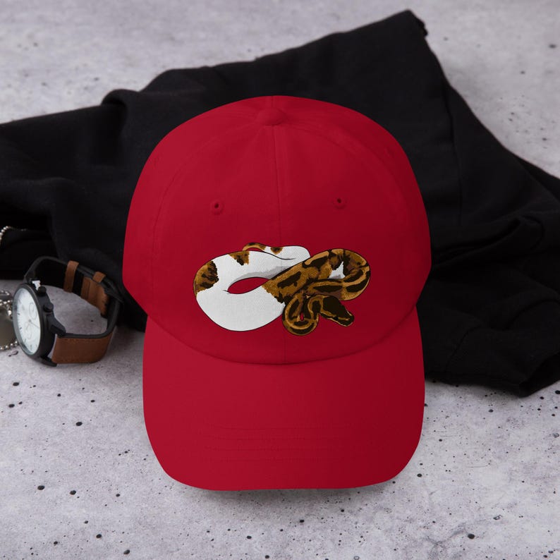 May include: A red baseball cap featuring a detailed illustration of a snake in shades of brown, white, and black. The cap is made of a soft material and has a curved brim. A black jacket and a watch with a brown strap are in the background.