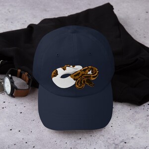 May include: Navy blue baseball cap featuring a graphic of a snake in white, brown, and tan. The cap is made of a soft material and has a curved brim. A watch with a brown leather band is visible in the background.