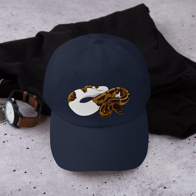 May include: Navy blue baseball cap featuring a graphic of a snake in white, brown, and tan. The cap is made of a soft material and has a curved brim. A watch with a brown leather band is visible in the background.