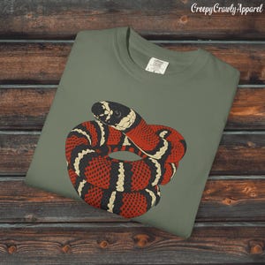 Puede incluir: Camiseta verde oliva con una ilustración de una serpiente enroscada. La serpiente tiene un patrón rojo, negro y crema. El texto "Creepy Crawly Apparel" está en la esquina superior derecha.