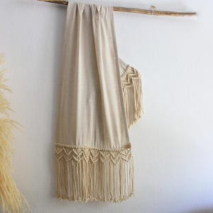 Op de afbeelding: Een beige macramé wandkleed met een geweven ontwerp en franjedetails. Het textiel is over een houten tak gedrapeerd, wat een natuurlijke en bohemien uitstraling creëert. De macramé heeft een geometrisch patroon en lange, vloeiende franjes.