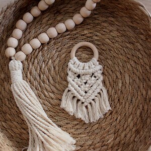 Op de afbeelding: Een crèmekleurige macramé babybijtring met een houten ring en franjes. Een houten kralen slinger met een kwast is ook aanwezig. De items worden gepresenteerd op een geweven mand.
