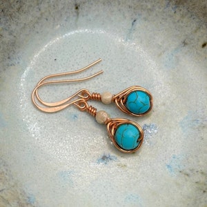 May include: A pair of turquoise and copper wire wrapped earrings with white beads. The earrings have a teardrop shape and are made with copper wire.