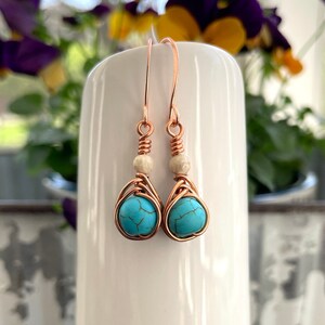 May include: Copper wire earrings with turquoise stones and white beads. The earrings are hanging from a white surface.