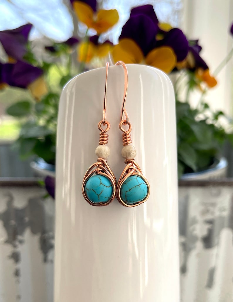 May include: Copper wire earrings with turquoise stones and white beads. The earrings are hanging from a white surface.