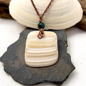 May include: A handcrafted pendant necklace featuring a square, cream-colored shell pendant with horizontal ridges. The pendant is accented with a teal bead and copper wire detailing, suspended from a copper chain. The necklace is displayed on a dark gray stone.