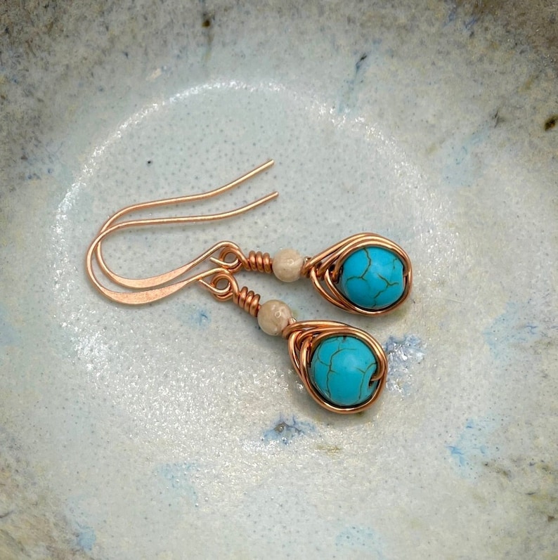 May include: A pair of turquoise and copper wire wrapped earrings with white beads. The earrings have a teardrop shape and are made with copper wire.