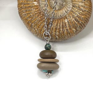 May include: A silver chain necklace featuring a pendant composed of a small green speckled bead, two smooth brown stone discs, a smaller turquoise bead, and a silver metal bead cap, all suspended in front of a large, circular, textured fossil.