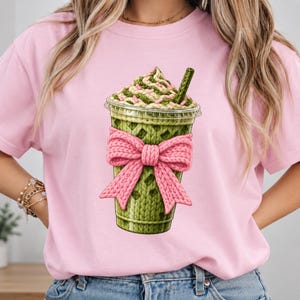 Pode incluir: Uma t-shirt rosa claro com um gráfico de uma bebida matcha verde num copo de plástico transparente. A bebida tem um aspeto de malha, coberta com natas batidas e granulado rosa, e decorada com um grande laço de malha cor-de-rosa.