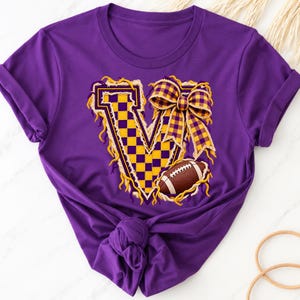 May include: A purple short-sleeved t-shirt featuring a large letter V with a yellow and purple chequered pattern. The letter is decorated with a yellow and purple plaid bow and a brown football graphic with white laces and stitching.