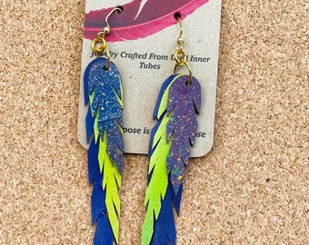 May include: A pair of blue and green feather earrings made from recycled inner tubes. The earrings are hanging from a brown card with the text "Consciously Crafted Jewelry" and "Jewelry Crafted From Used Inner Tubes".