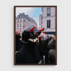Peut inclure: Photographie encadrée d'une personne dans une foule, portant un casque noir avec des cornes rouges et une visière dorée. La personne est vêtue de noir, avec un arrière-plan flou de bâtiments et d'autres personnes.
