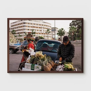 May include: A street vendor prepares fruit, including pineapples, near a black car. A person in traditional attire stands nearby. The scene is set against a backdrop of buildings and palm trees, suggesting an urban market.