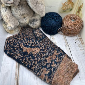May include: A navy blue and brown hand-knitted mitten with a deer pattern, beside two balls of yarn and a stuffed bunny. The mitten is made of wool and has a detailed design. A children's book with rabbit illustrations is in the background.