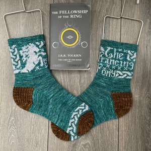 May include: Two teal blue knitted socks with white and brown accents. One sock features a white design of a creature with a crown, the other sock has the text "The Francing Pony" in white. The socks are hanging on metal hangers.