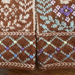 May include: Close-up of two sections of a knitted textile. The brown fabric features intricate patterns in cream, light blue, purple, and green. The design includes geometric shapes and floral motifs, with a decorative border.