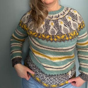 May include: A teal and gray striped sweater with a yellow and gray patterned yoke. The yoke features a design of stylized birds.