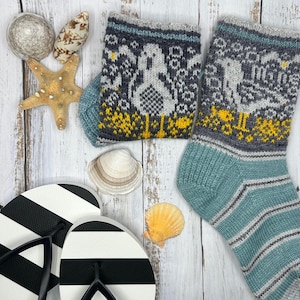 以下が含まれることがあります： 白と黄色のカモメの模様がついた、ターコイズブルーのニットソックスのペア。ソックスは、貝殻とヒトデのある白い木製の背景の上に置かれています。