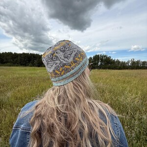 Puede incluir: Un gorro de punto gris con un patr&oacute;n geom&eacute;trico en amarillo y blanco, y un borde azul claro. El gorro se lleva al aire libre en un campo de hierba alta bajo un cielo nublado. La persona que lleva el gorro est&aacute; de espaldas a la c&aacute;mara.