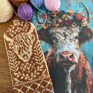 May include: A brown and cream-colored knitted mitten with a geometric pattern, next to a postcard featuring a cow with a floral crown. Several balls of yarn in various colors, including brown, blue, and pink, are arranged around the mitten and postcard.