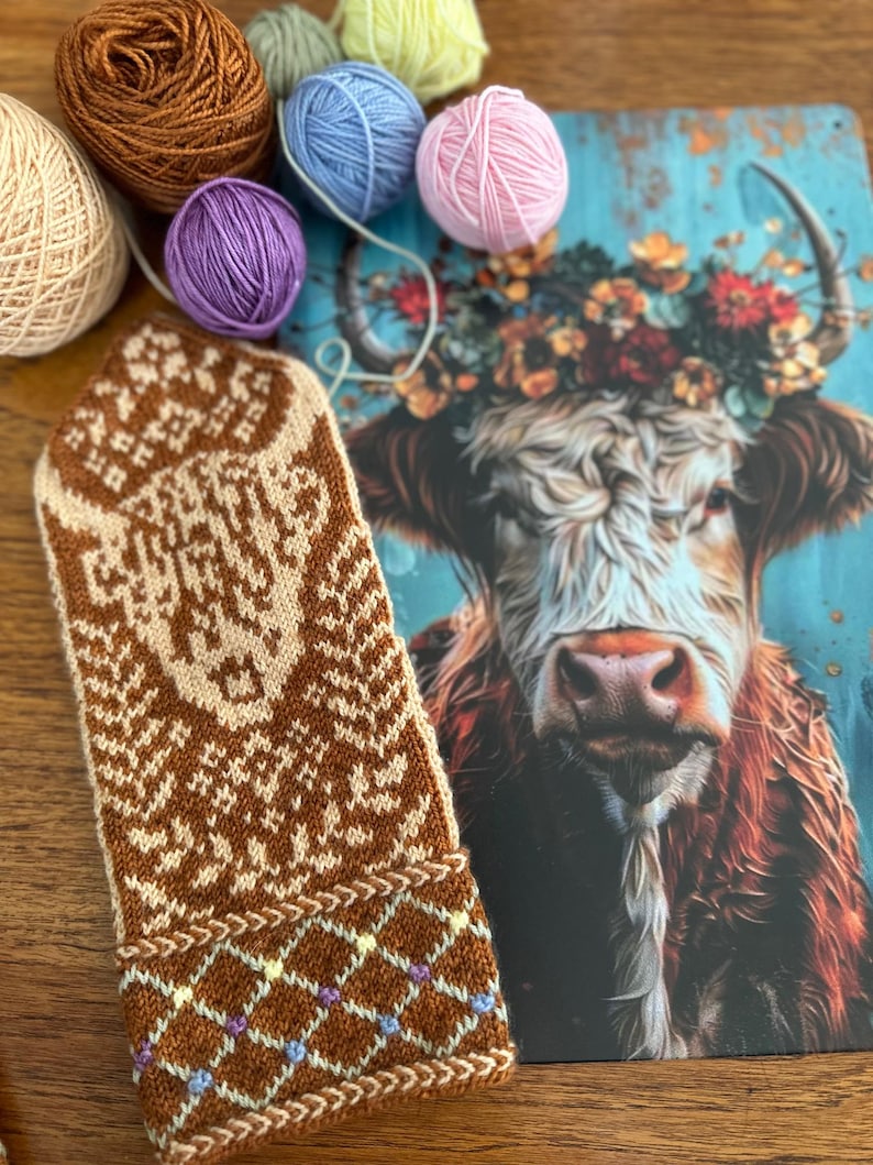 May include: A brown and cream-colored knitted mitten with a geometric pattern, next to a postcard featuring a cow with a floral crown. Several balls of yarn in various colors, including brown, blue, and pink, are arranged around the mitten and postcard.