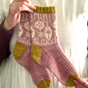 May include: A pair of knitted socks with a floral pattern. The socks have a pink body with cream floral designs, mustard yellow heels and toes, and a mustard yellow ribbed cuff. The socks are held up by a person wearing a white shirt.