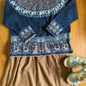 Può includere: Un maglione blu navy con un motivo dettagliato in bianco e marrone, abbinato a una gonna marrone e sandali turchesi. Il maglione presenta uno scollo rotondo e maniche lunghe con motivi coordinati. La gonna è di colore neutro.