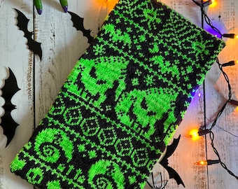 May include: A black and neon green knitted cowl with a Halloween theme. The cowl features a repeating pattern of skulls and other spooky shapes. Decorative bats, a spider, and a green hand with purple nails are also in the image.