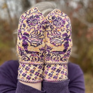 Puede incluir: Un par de mitones de punto con un patrón complejo en tonos morado, beige y naranja. Los puños tienen un diseño floral repetido y están ribeteados con un hilo contrastante. Los mitones se sostienen frente a una persona.