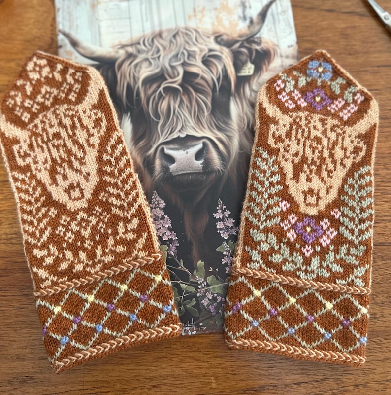 May include: A pair of brown and tan knitted mittens with intricate patterns. The mittens feature a diamond pattern on the cuffs and floral designs on the upper portion. A postcard with a Highland cow is in the background.