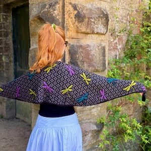 Könnte beinhalten: Ein dunkelgraues Stricktuch mit einem sich wiederholenden geometrischen Muster und verstreuten Libellenmotiven in leuchtendem Limettengrün, Lila und Türkis. Das Tuch liegt über den Schultern und zeigt die komplexe Strickstruktur.