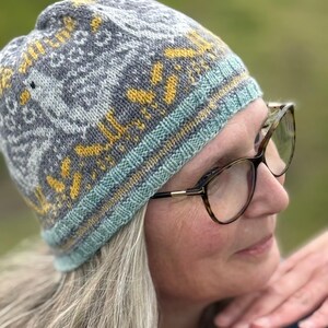 Puede incluir: Un gorro de punto gris con un patr&oacute;n geom&eacute;trico en amarillo y azul claro. El gorro tiene un borde acanalado en azul claro y amarillo. El gorro lo lleva una persona con gafas.