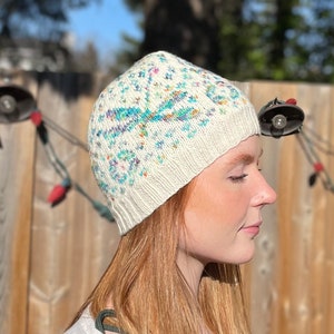 May include: A white knitted hat with a colorful floral pattern. The hat is worn by a person with long, red hair.