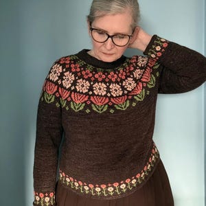 May include: A dark brown long-sleeved sweater featuring a round neckline and a colorful floral pattern around the yoke, cuffs, and hem. The knit design includes stylized flowers in shades of pink, light green, and white against the dark base.