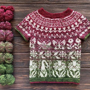 May include: A short-sleeved, crew-neck sweater in burgundy, white, and green, with a floral pattern. The sweater is displayed next to several balls of yarn in coordinating colours. The yarn and sweater are on a dark wood surface.