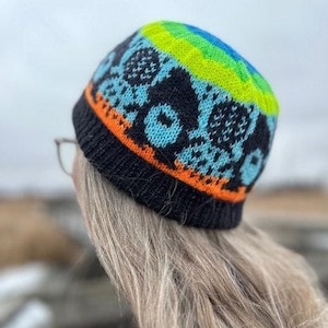 Könnte beinhalten: Eine gestrickte Beanie-Mütze mit einem farbenfrohen Design. Die Mütze hat eine schwarze Basis mit einem orangefarbenen Band am unteren Rand. Das Hauptmuster umfasst blaue und weiße geometrische Formen, mit einem oberen Abschnitt aus blauen, grünen und gelben Streifen.