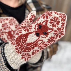 Op de afbeelding: Een paar gebreide wanten in crème en rood. De wanten hebben een rendierontwerp en sneeuwvlokpatronen. De manchetten hebben een neutraal kleur met een ruitpatroon. De wanten zijn ontworpen voor koud weer.