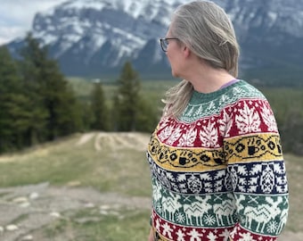 May include: A colorful sweater with a mountain-themed design. The sweater features a pattern of trees, snowflakes, and animals in red, white, green, yellow, and navy blue. The person is wearing glasses and looking at a mountain range.