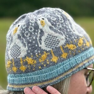 Puede incluir: Un gorro de punto gris, azul y amarillo, con un dise&ntilde;o de gansos. El gorro tiene un borde azul con detalles amarillos y un patr&oacute;n repetido de gansos en blanco y amarillo. El gorro est&aacute; hecho de hilo suave.