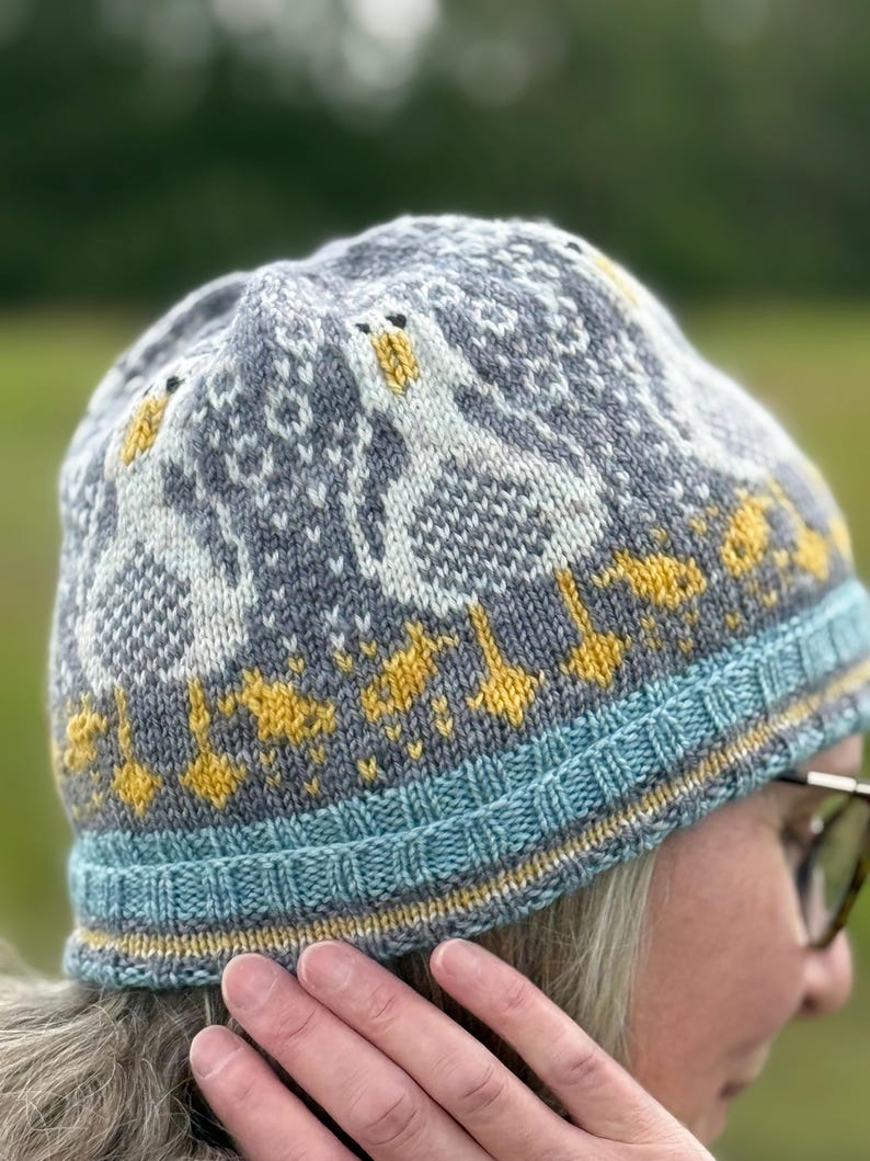 Puede incluir: Un gorro de punto gris, azul y amarillo, con un dise&ntilde;o de gansos. El gorro tiene un borde azul con detalles amarillos y un patr&oacute;n repetido de gansos en blanco y amarillo. El gorro est&aacute; hecho de hilo suave.