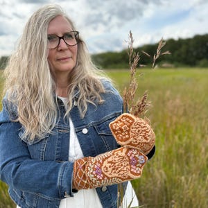 May include: A person wearing a denim jacket and holding a pair of knitted mittens. The mittens feature a brown, tan, and purple floral design. The background is a field of tall grass under a cloudy sky.